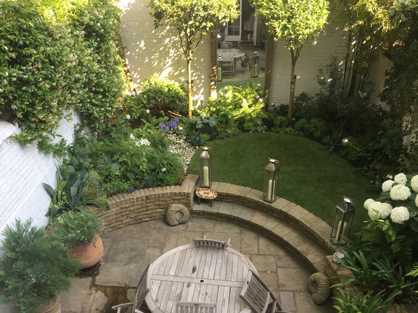 A garden with a round wooden table, chairs, and various plants.